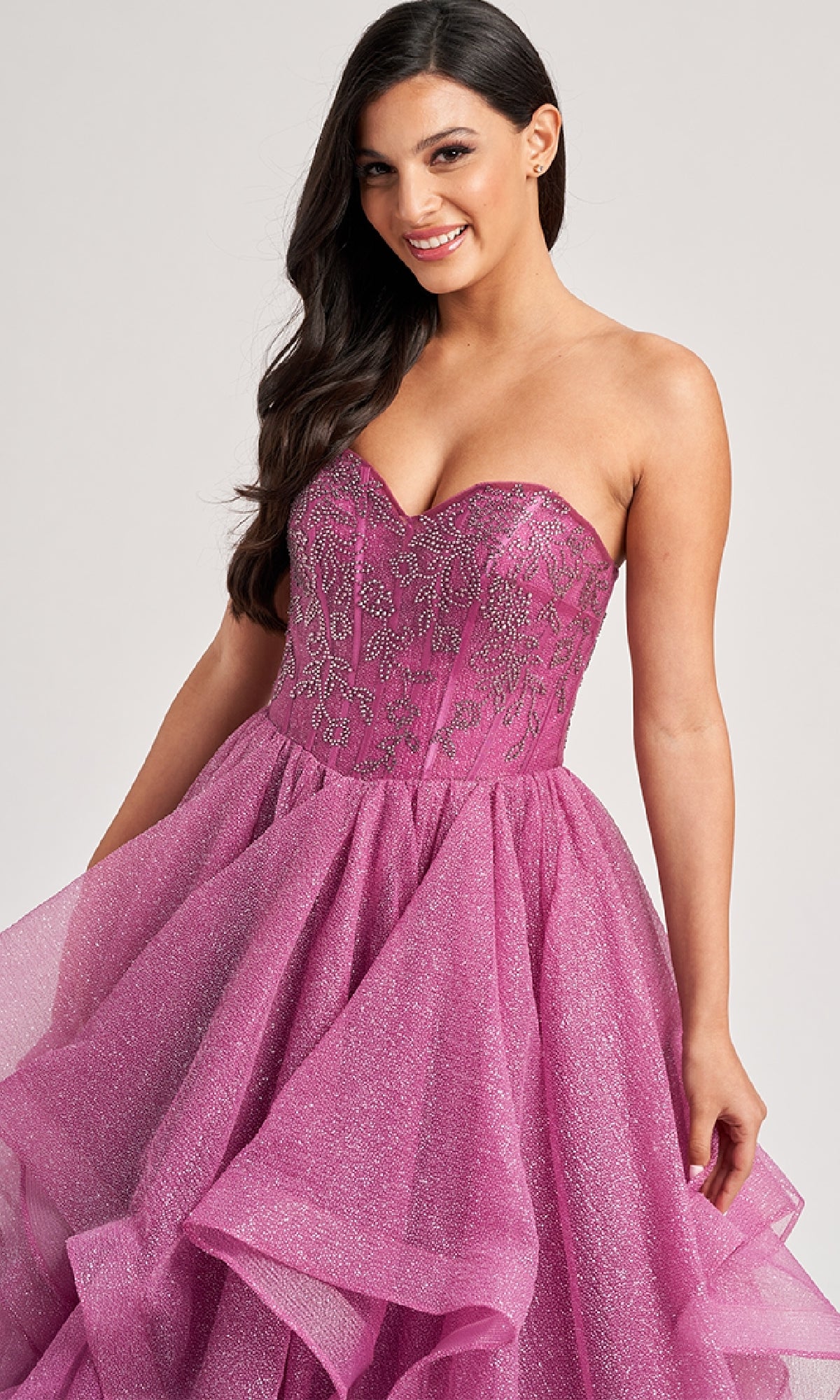 A woman stands smiling at the camera in the Colette CL8200 long prom ball gown, featuring a strapless beaded corset bodice and a ruffle-tiered glitter tulle skirt.